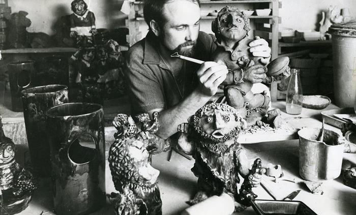 The black and white photo shows John Hughes sculpting a clay rugby Grogg, surrounded by various miscellaneous characters and pottery.